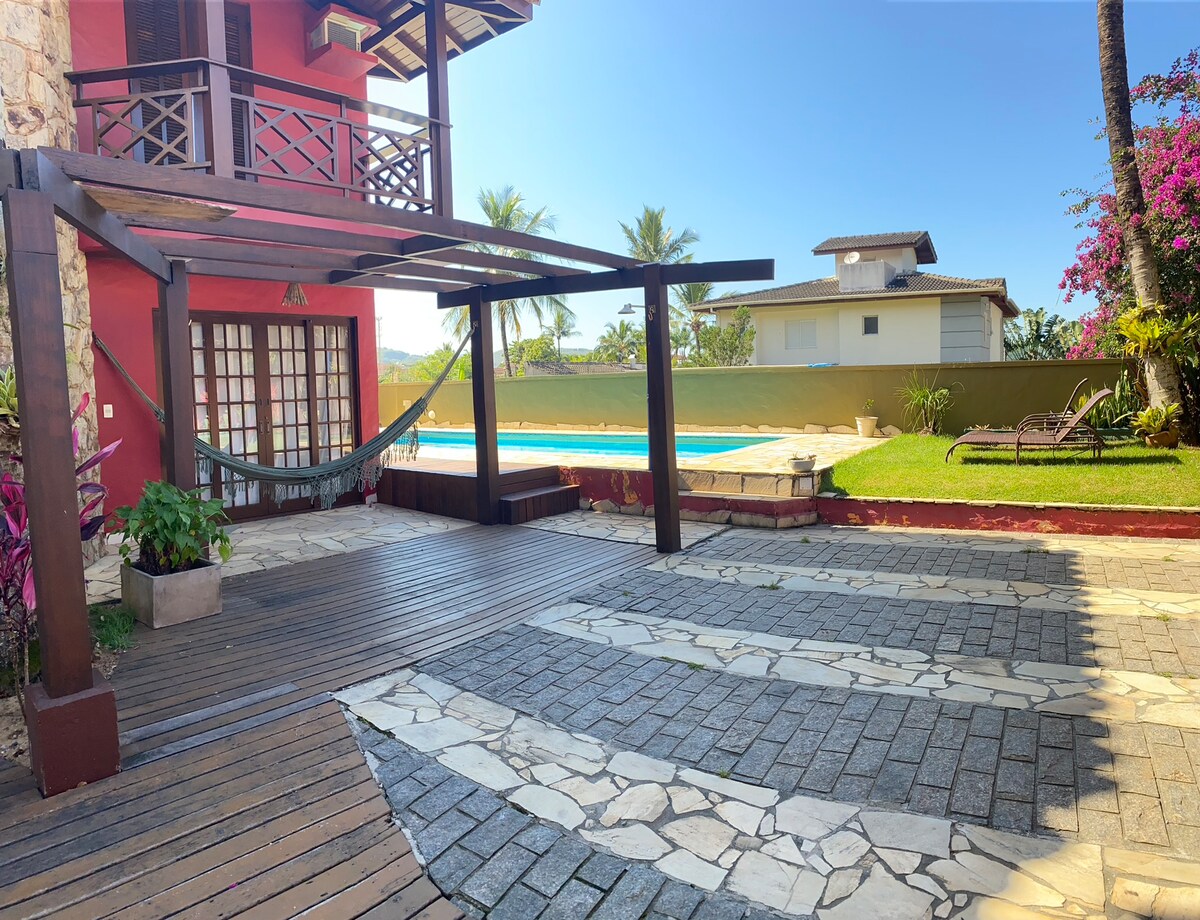 An outdoor lounge area features a wooden deck with a hammock hanging beneath a covered structure. Colorful plants surround the space, while a pool is visible in the background, complemented by palm trees and a clear blue sky.