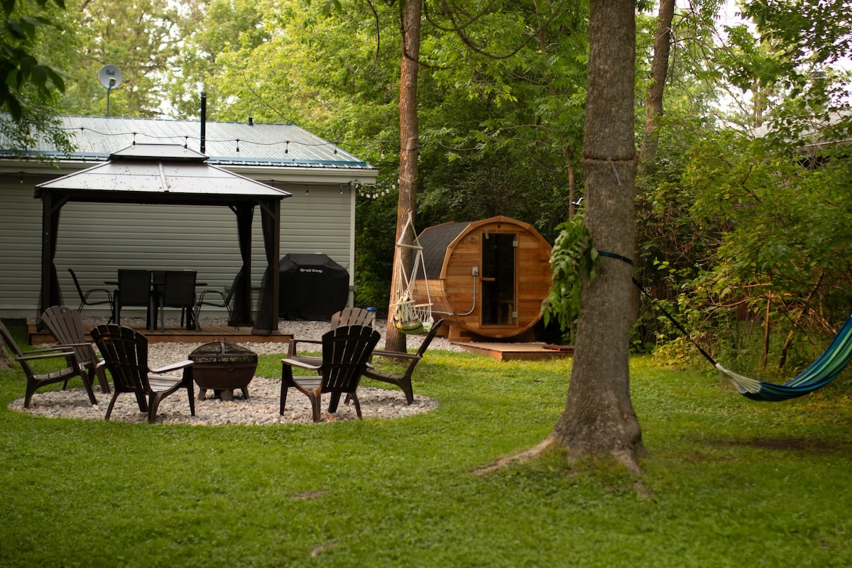 A cozy outdoor area features a fire pit surrounded by several wooden chairs on a grassy lawn. Nearby, a swing hangs from a tree, and an outdoor cedar sauna is visible alongside a structured gazebo with a dining table and grill.
