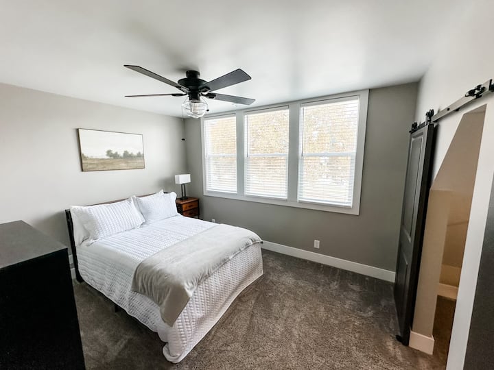 Entra en nuestro cómodo dormitorio del segundo piso con una cama de tamaño completo, un armario espacioso, un vestidor y una mesita de noche equipada con una lámpara que incluye puertos de carga. Disfruta de la vista de la vibrante calle principal y el parque, mostrando el encantador vecindario.