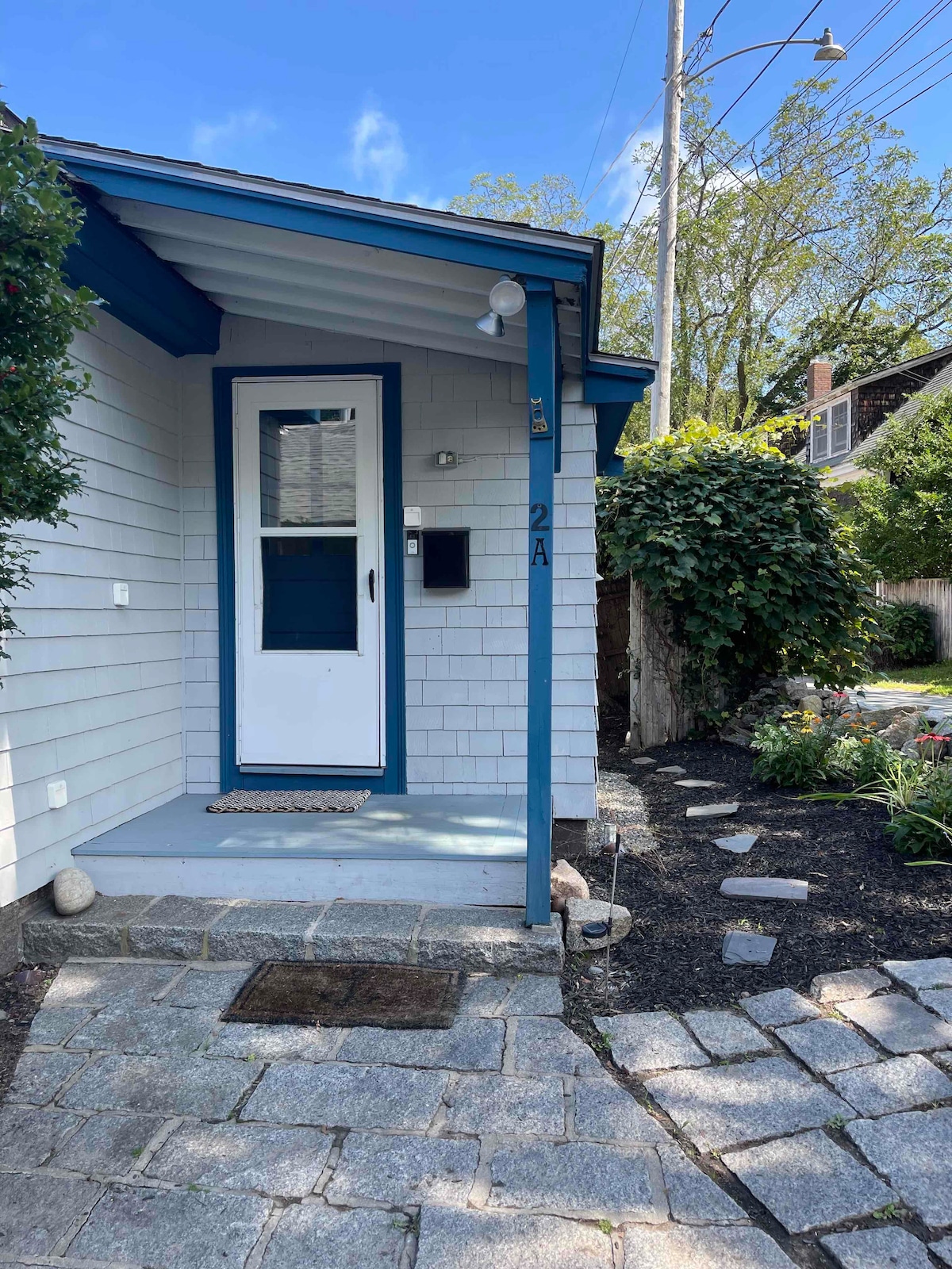 The entrance of the cottage is highlighted by a charming blue door and a comfortable porch area. Surrounding pathways feature stone paving and well-maintained landscaping, including a mixture of shrubs and flowering plants, enhancing the inviting approach to the home.