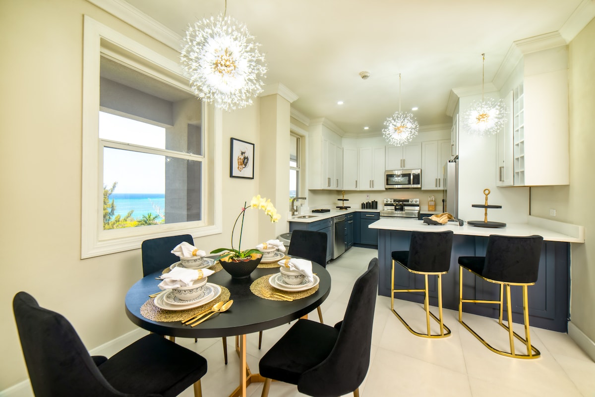 A modern dining area features a sleek round table set for six, adorned with elegant tableware. Stylish black chairs with gold accents surround the table. A bright window offers a glimpse of ocean views, while contemporary light fixtures hang above, enhancing the space.