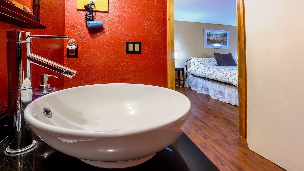 A modern sink with a rounded design is featured in the foreground, accompanied by a sleek faucet. The rich red wall adds a vibrant contrast. A doorway leads to a cozy bedroom with a floral-patterned bedspread, seen in the background.