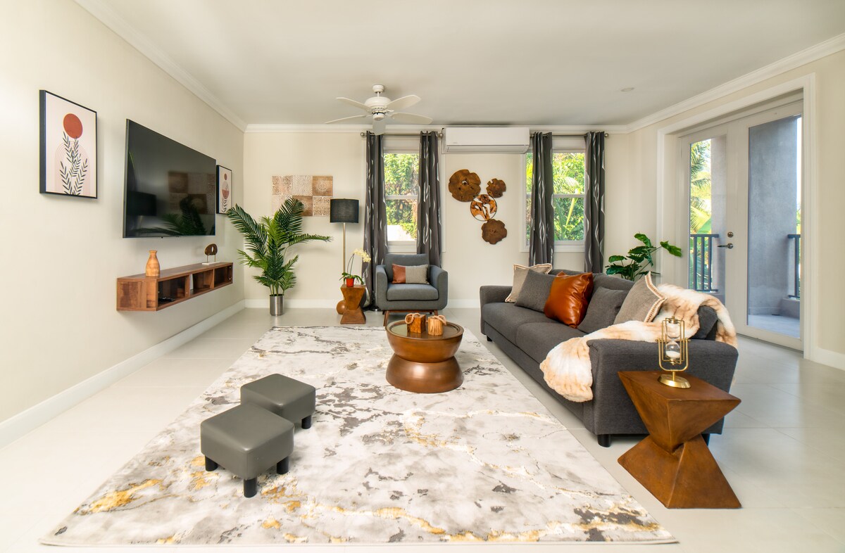 A modern living area features a comfortable gray sofa and armchair, complemented by a round wooden coffee table. Decorative art pieces add interest to the walls, and large windows provide natural light, creating a bright and inviting space. Lush greenery can be seen through the glass doors.