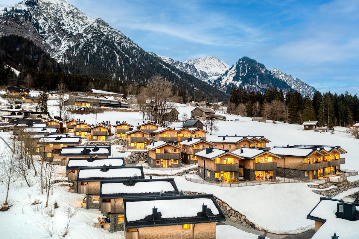 The image captures a snow-covered landscape dotted with charming chalets, each warmly illuminated from within. Majestic mountains rise in the background under a clear sky, showcasing a serene winter setting that invites relaxation and exploration.