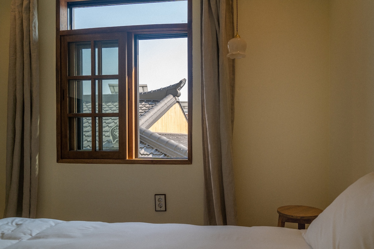 A bright room is displayed with a spacious bed and a window featuring wooden frames. Natural light is allowed in, illuminating the soft tones of the walls. A small wooden stool is placed nearby, complementing the warm atmosphere of the space.