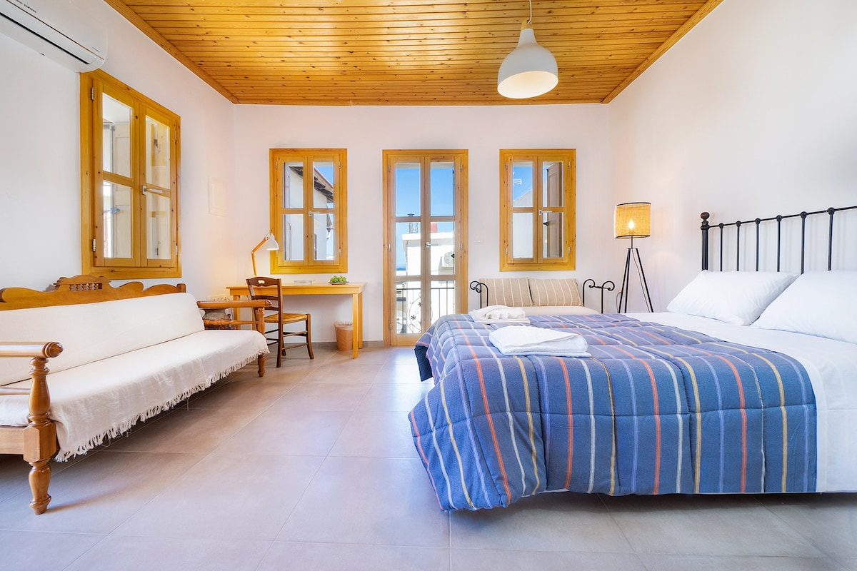 A spacious bedroom features a double bed with a blue striped blanket and natural light pouring through large windows. A cozy sofa and a desk with a lamp are positioned to one side, while a sleek floor lamp and wooden ceiling add to the room's charm.