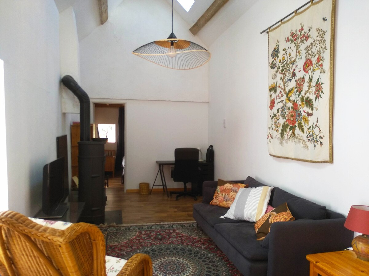 A spacious living area is featured, adorned with a rustic wood-burning stove and a large window providing natural light. A comfortable sofa is positioned alongside a woven chair, complemented by a decorative wall hanging and a workspace with a desk. The flooring is wooden and enhances the room's warmth.