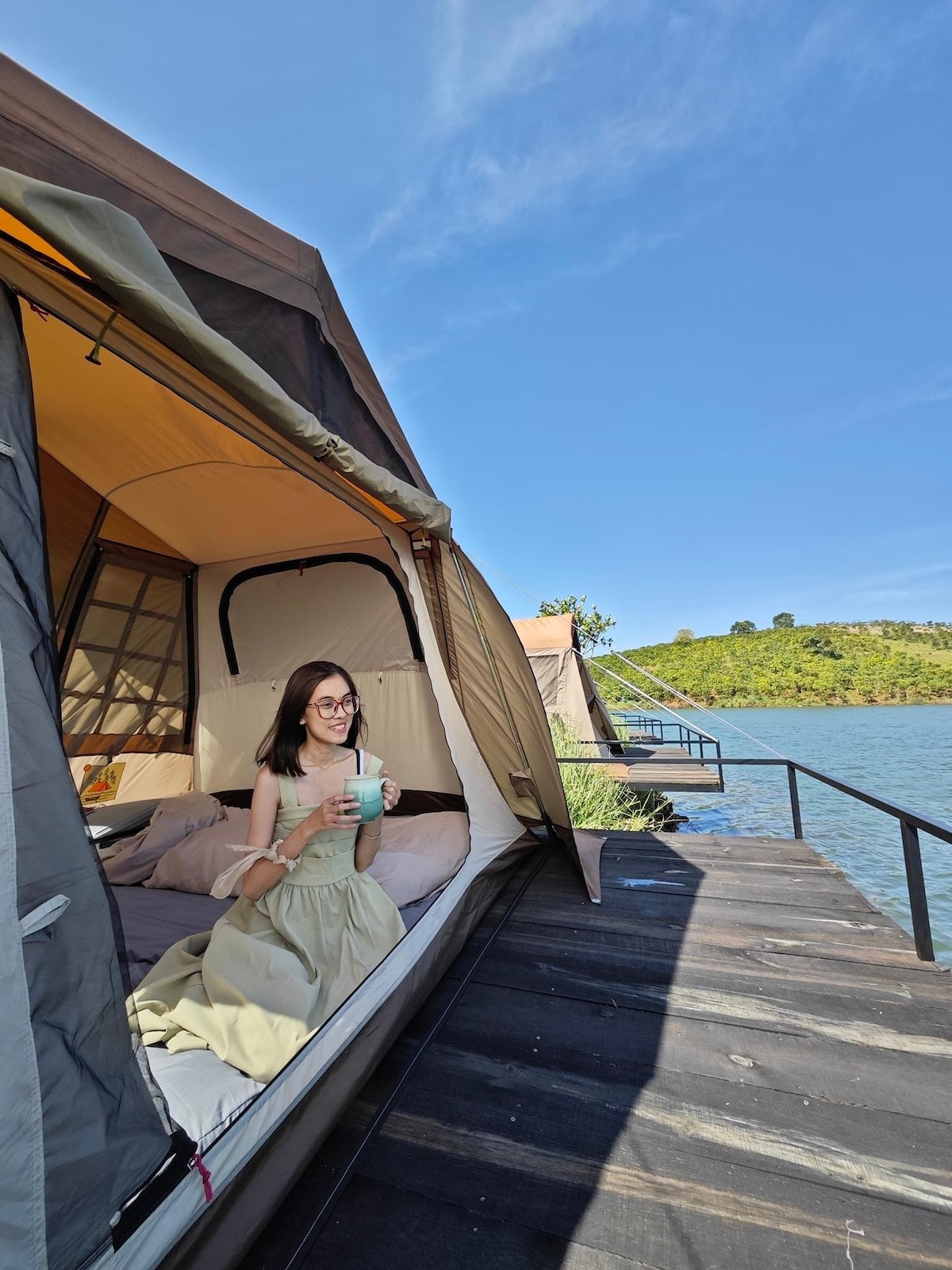 A comfortable glamping tent is positioned close to the water's edge, featuring a spacious interior with a soft bed. Natural light floods the tent through a large opening, creating a calm ambiance. A scenic view of the lake and surrounding greenery is visible from the deck.