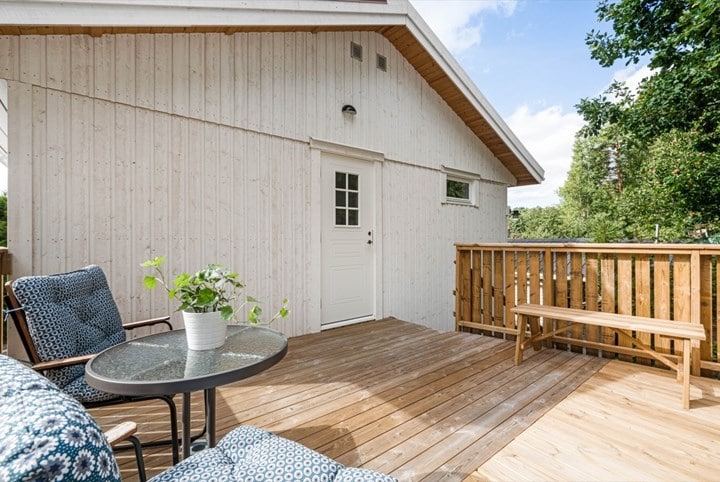 A wooden deck features a small round table with a potted plant, accompanied by two cushioned chairs in a patterned fabric. A wooden bench sits alongside the railing, providing an open view of the surrounding greenery.