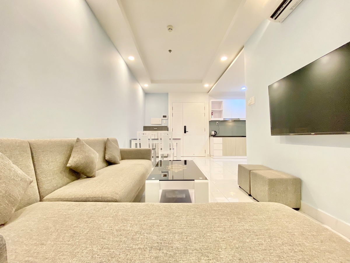 A modern living area features a light blue wall and polished white flooring. A spacious gray sofa and matching ottoman are positioned around a glass coffee table. A wall-mounted television and a well-equipped kitchen area are visible in the background, enhancing the open and airy feel.