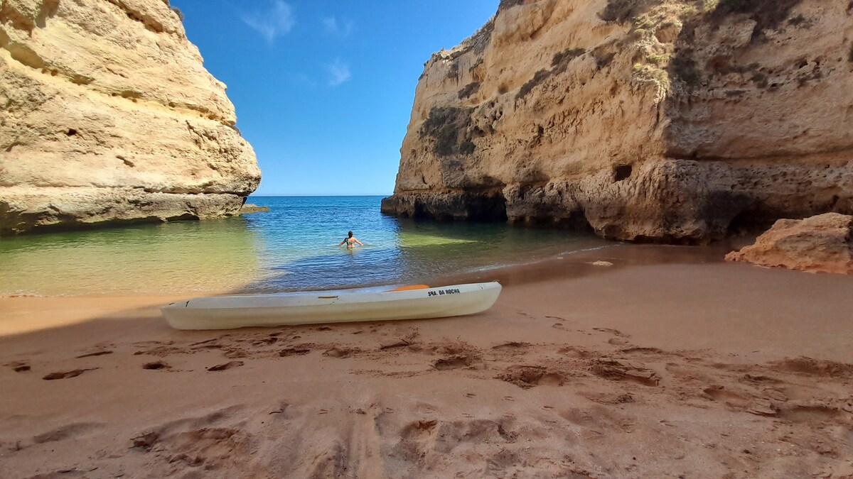 A tranquil beach scene showcases golden sand bordered by rugged cliffs. Calm turquoise waters reflect blue skies, while a kayak is positioned on the shore. A figure can be seen wading into the water, creating a serene atmosphere in a secluded coastal setting.
