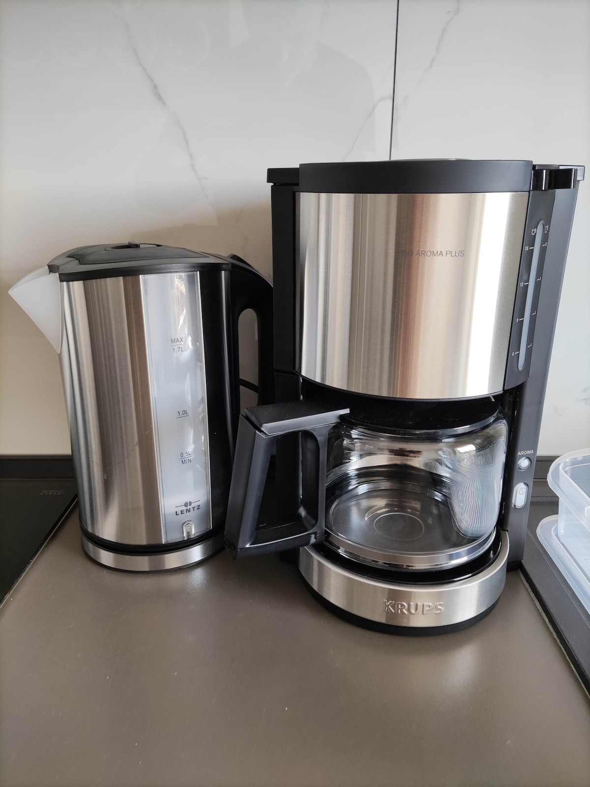 A modern coffee setup is featured, consisting of a sleek stainless steel kettle and a coffee maker. The coffee maker has a transparent glass carafe, allowing visibility of the brewed coffee inside. Both appliances are placed on a smooth countertop, enhancing the kitchen's contemporary look.