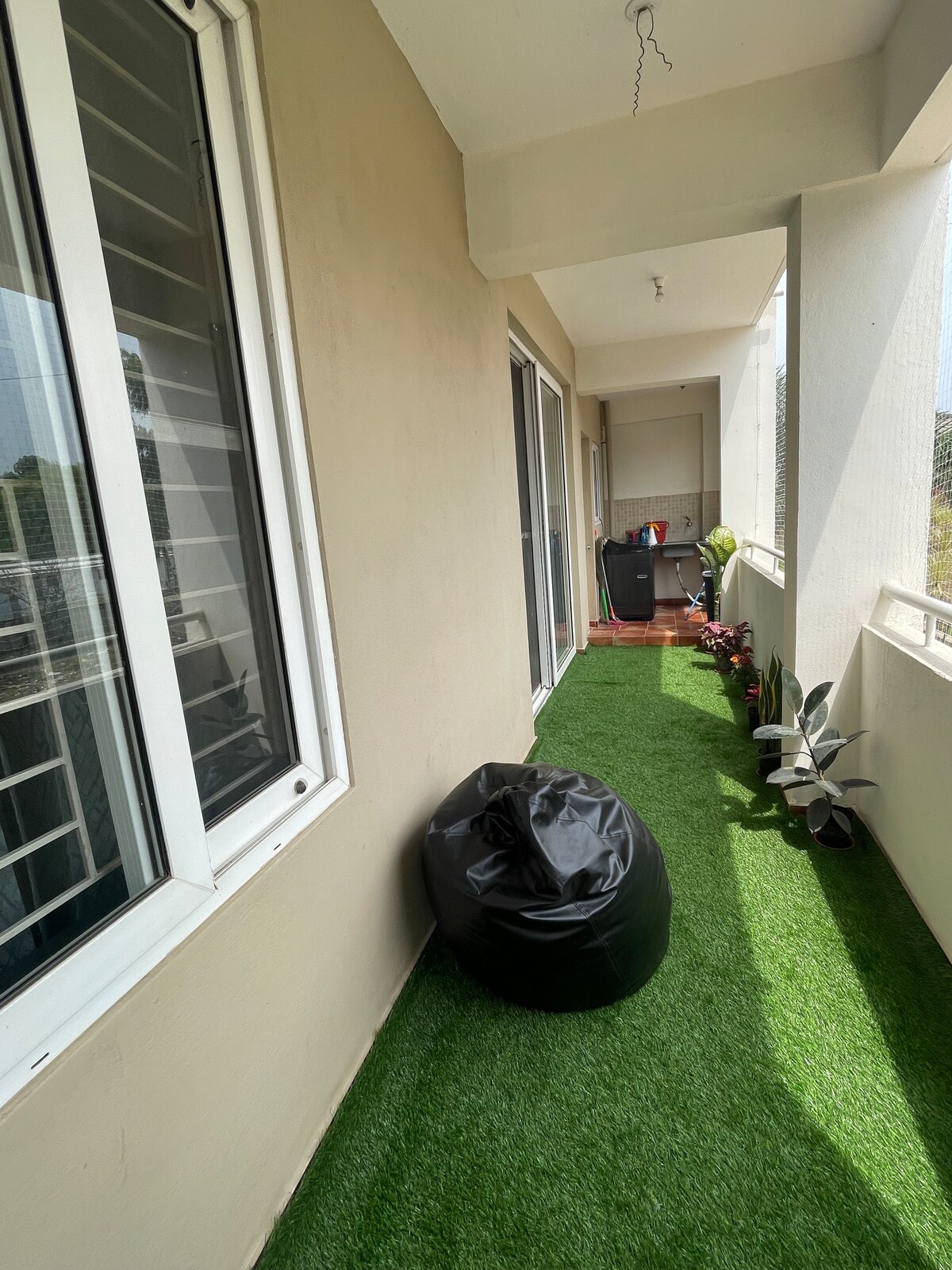 A balcony area is shown featuring a synthetic lawn, creating a green space. A black bean bag is positioned along the side, and potted plants line the edge. Large windows offer views of the outside while allowing natural light into the area.