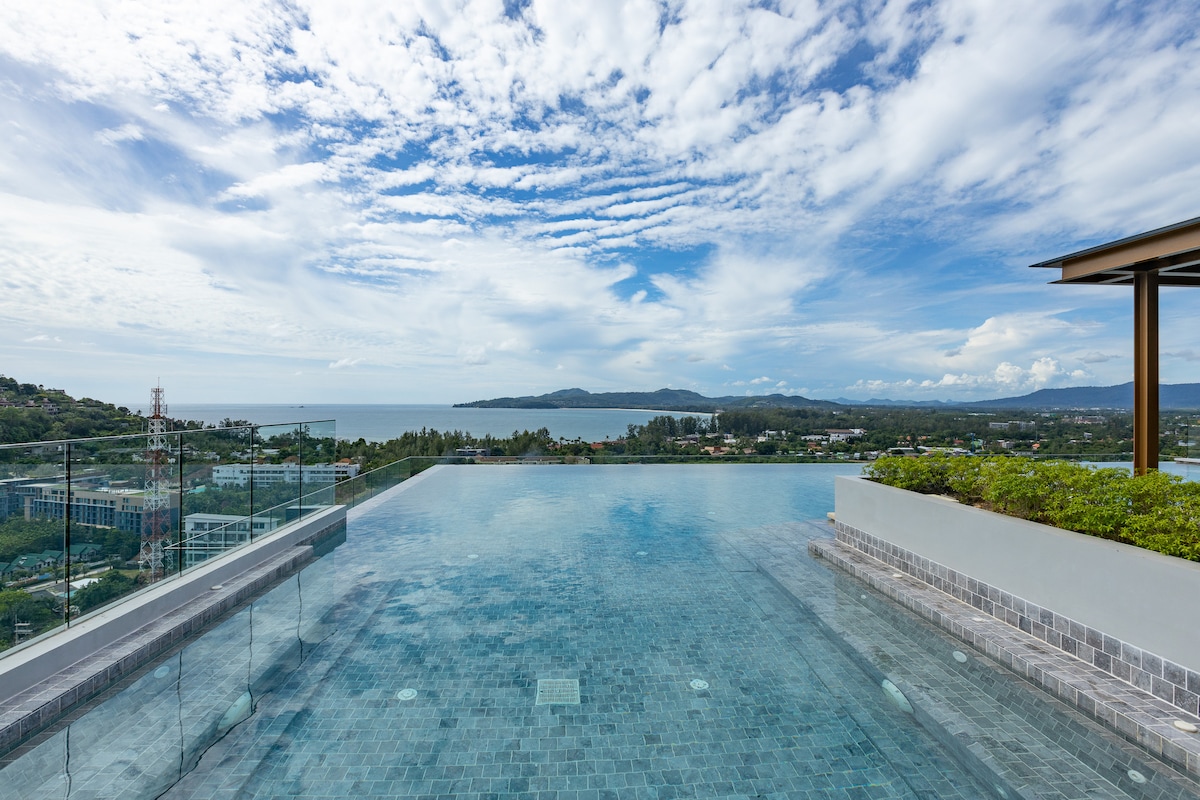 A serene rooftop infinity pool overlooks the coastline, blending with the sky above. Lush greenery is visible along the pool's edge, contributing to a refreshing ambiance. The horizon showcases distant hills and the sparkling sea under a partly cloudy sky.