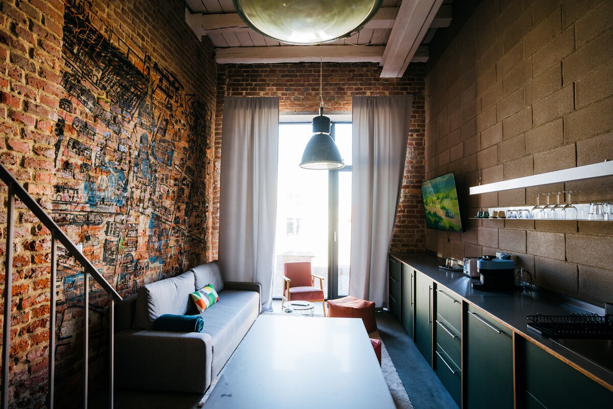 A spacious living area features exposed brick walls and a large window draped with sheer curtains, allowing natural light to fill the room. A sleek kitchen with modern cabinetry and a flat-screen TV is present. A comfortable sofa and armchair provide seating options.