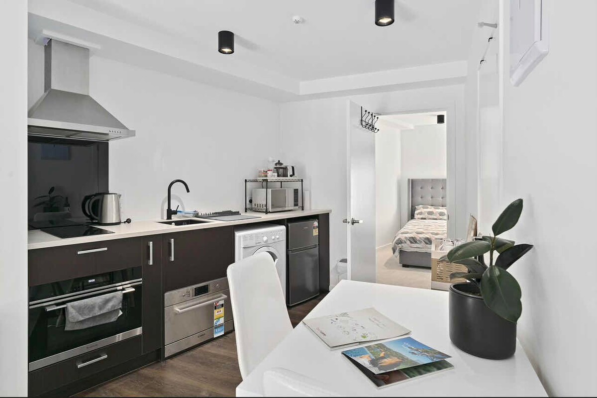 A modern kitchen features sleek black cabinetry and stainless steel appliances, including an oven, microwave, and kettle. Nearby, a small dining table is adorned with a potted plant and a brochure. The living area is visible through an open doorway, showcasing a comfortable bedroom layout.