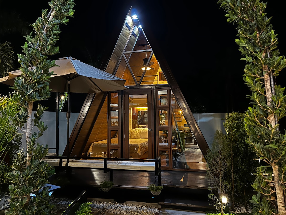 An A-frame cabin is illuminated at night, showcasing its inviting wooden structure and large glass doors. Lush greenery surrounds the exterior, and an umbrella provides shade over a seating area. Warm light glows from within, hinting at a cozy interior.