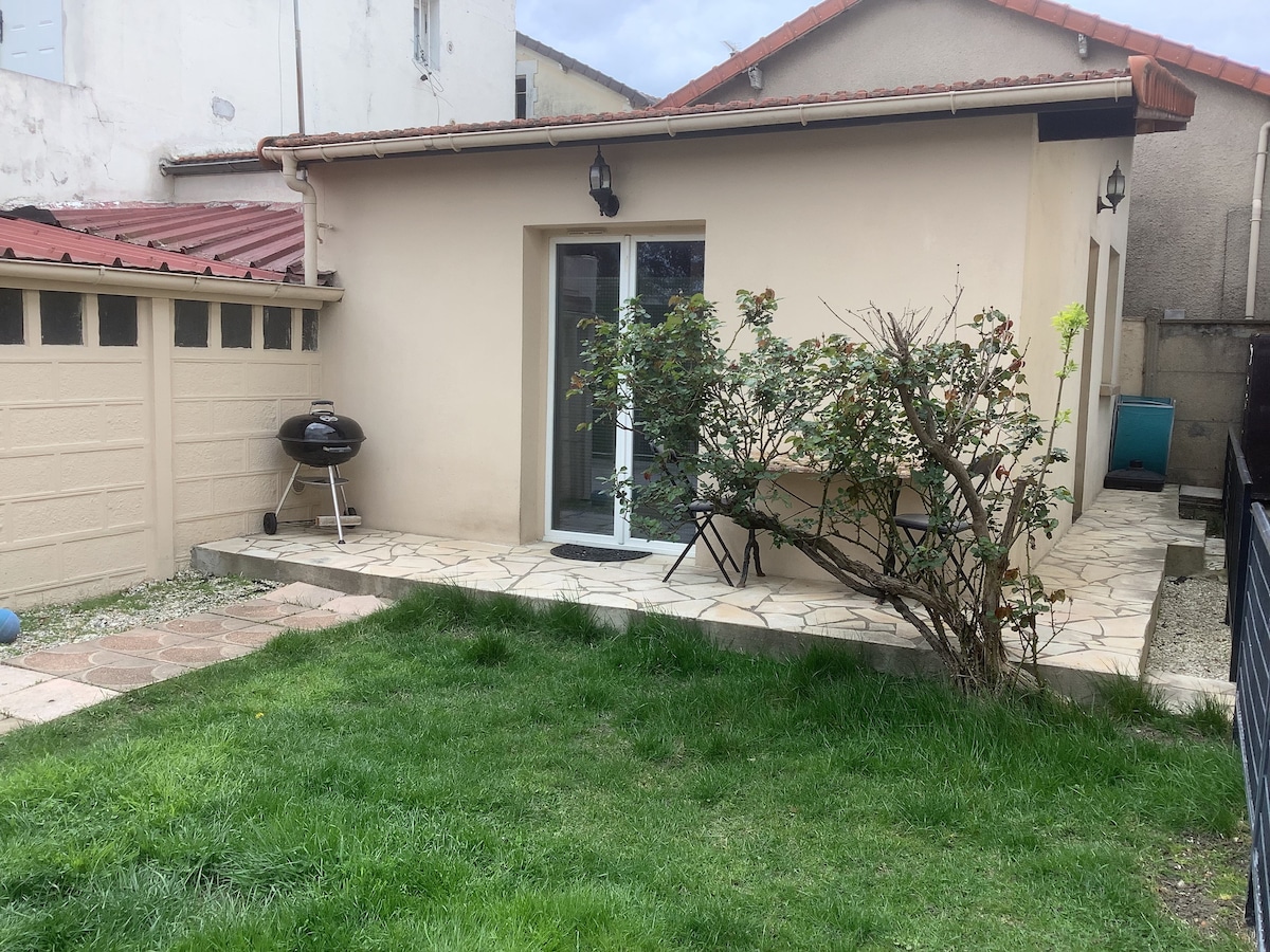 A small patio area is visible, featuring a grassy lawn bordered by a stone path. A barbecue grill is positioned nearby, alongside a metal chair and a small table. The entrance to the residence is framed by a glass door, surrounded by a few bushes.