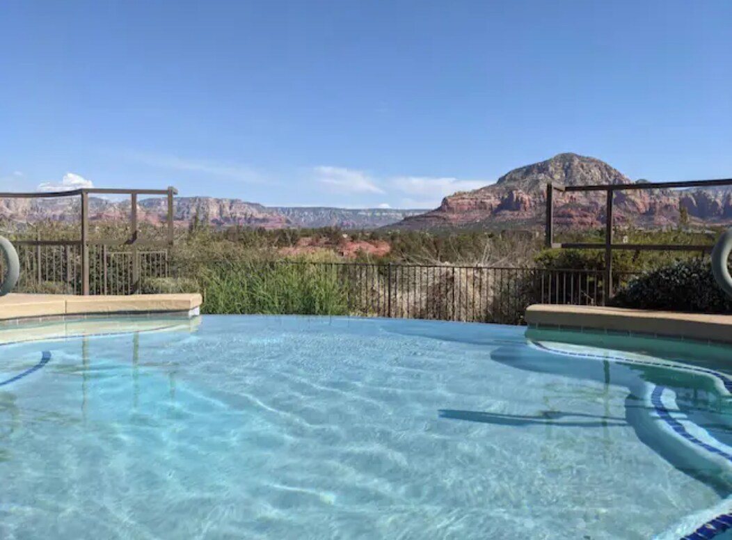 An inviting pool offers a refreshing place to relax, featuring clear blue water that reflects the surrounding landscape. The view showcases the natural red rock formations of Sedona under a bright sky, with a protective railing visible around the pool's edge.
