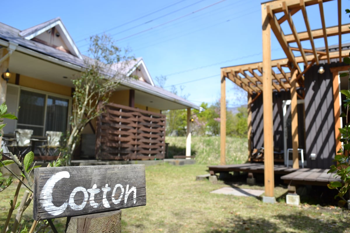 The exterior of the cottage is surrounded by a well-maintained lawn, with a wooden sign labeled 'Cotton' marking the entrance. A covered terrace is visible, complemented by seating arrangements. The architecture features a blend of traditional and modern elements, framed by natural greenery.