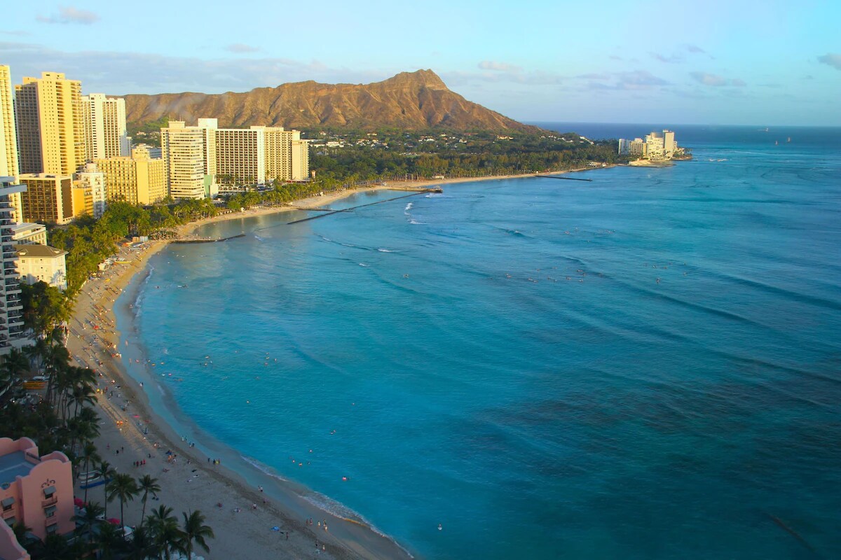 A sweeping view of Waikiki Beach is presented, showcasing calm blue waters gently lapping against a sandy shoreline. The backdrop features lush green mountains and a modern city skyline, creating a picturesque coastal scene under a blue sky.