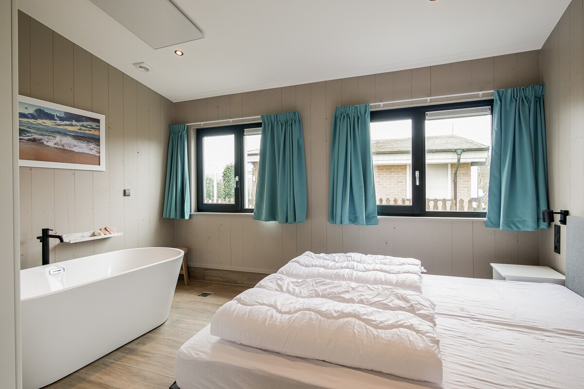 A serene sleeping area is designed with large windows allowing natural light to illuminate the space. A spacious bed is complemented by light bedding, while a freestanding bathtub is positioned nearby. Soft blue curtains frame the windows, enhancing the calm atmosphere.