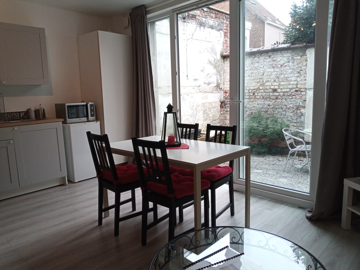A dining area features a rectangular table surrounded by four black chairs with red cushions. A large glass door provides a view of the outdoor courtyard, where a small bistro set is visible against the textured stone wall.