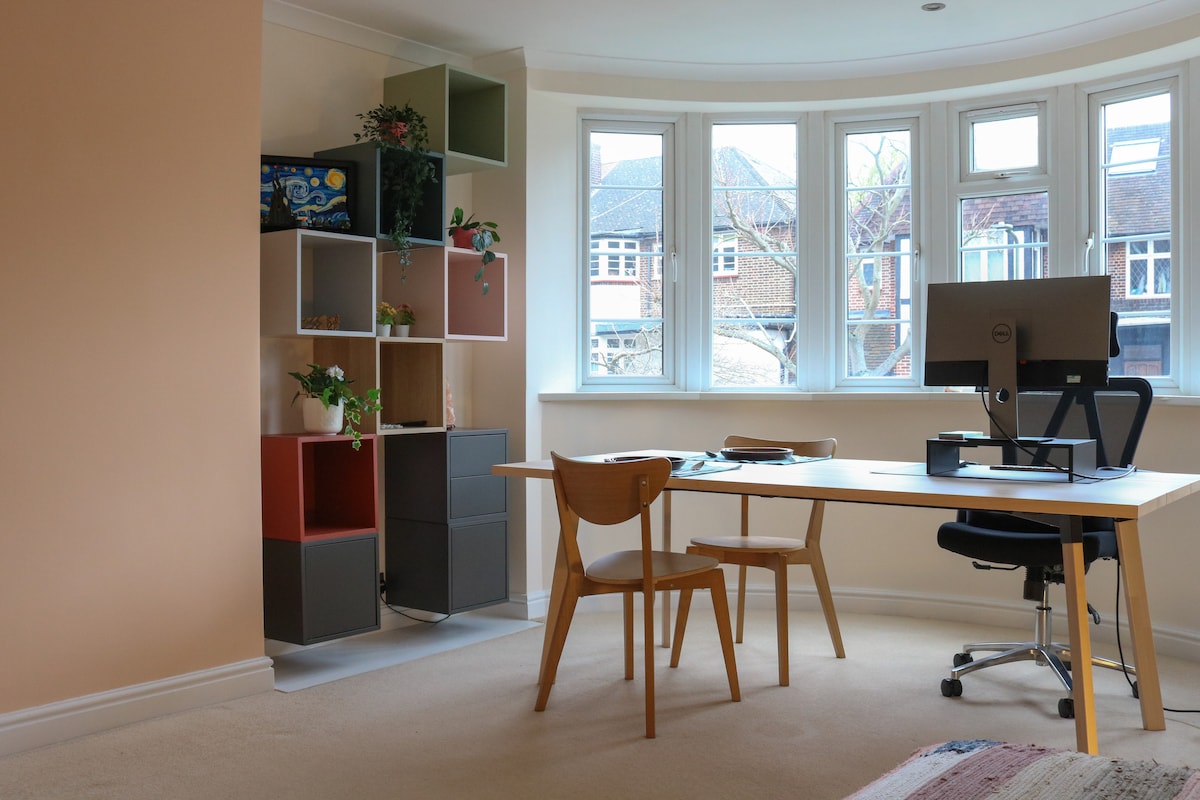A workspace is featured in a sunlit corner of the room, with a desk positioned near a bay window. A comfortable chair accompanies the desk, and colorful shelving units are arranged nearby, displaying plants and decor items.