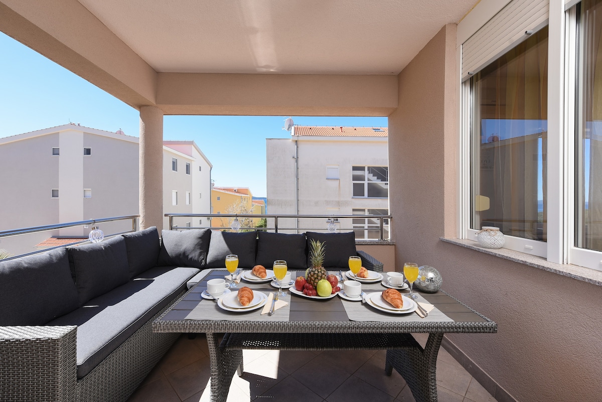 A spacious terrace is furnished with a large table surrounded by comfortable seating and is adorned with a neatly arranged breakfast spread featuring pastries, fruits, and drinks. The backdrop includes the neighboring buildings and glimpses of the serene sky.