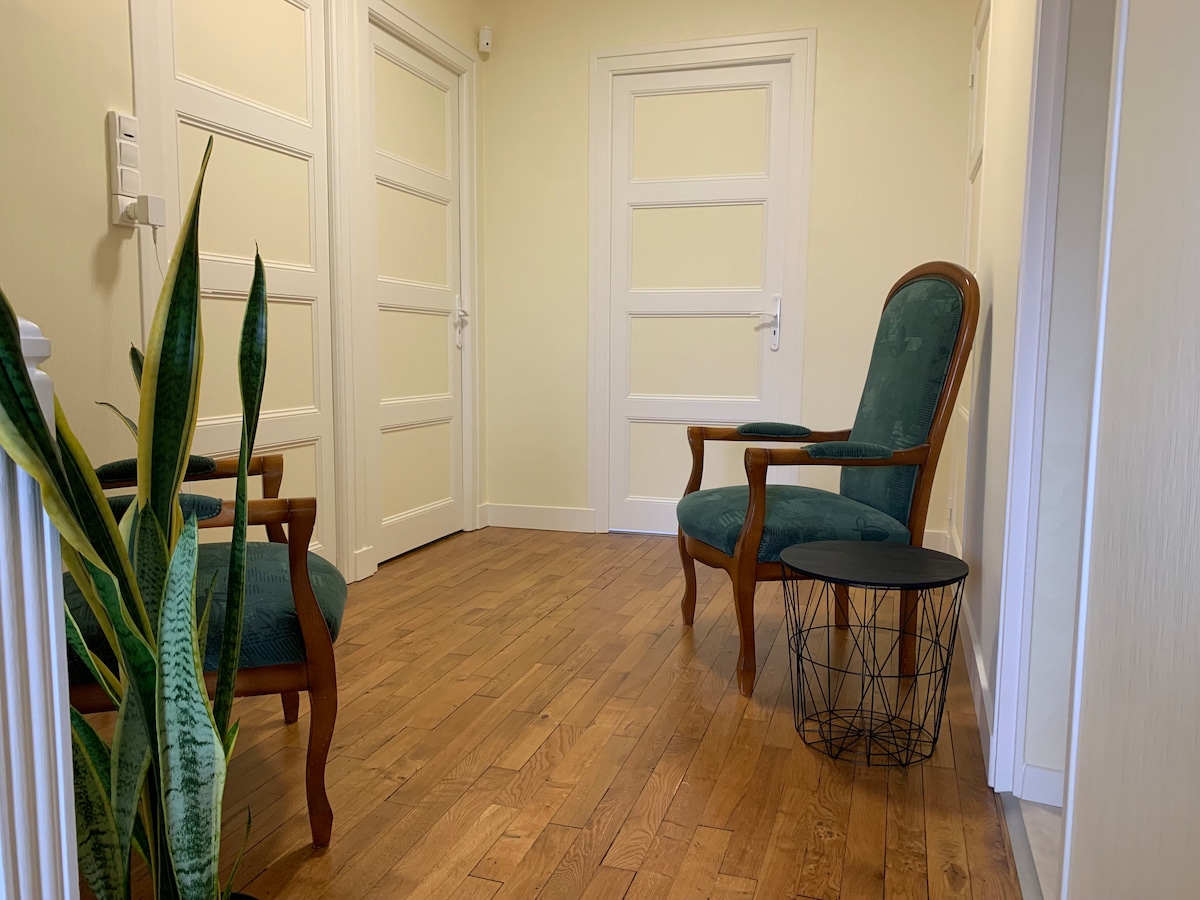 A hallway features light-colored walls and warm wooden floors. Two green upholstered chairs are positioned next to a small black side table. A tall plant is placed in the corner, adding a touch of greenery to the space.