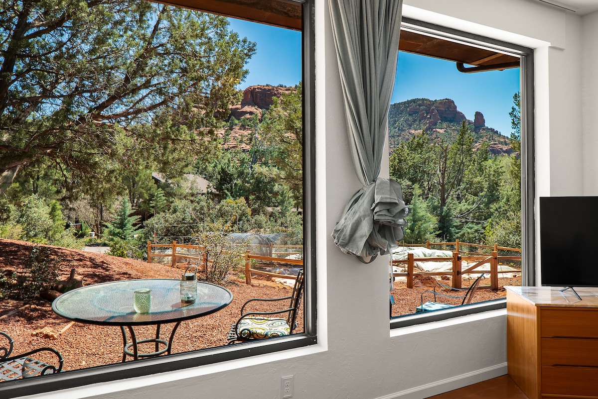 Large windows frame stunning views of red rock formations and lush greenery. A small round table with chairs rests on a private patio, inviting outdoor relaxation. Natural light fills the inviting living space, enhancing the connection with the serene outdoor environment.