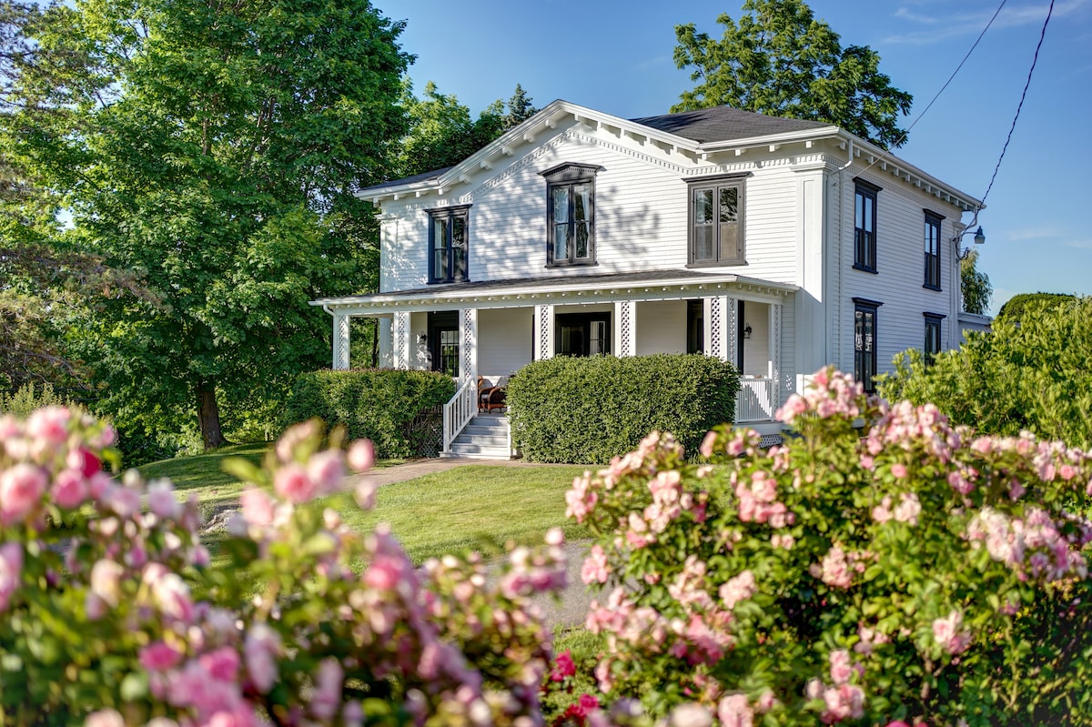 The exterior of the Borden House is shown, featuring a classic white facade with large windows and a welcoming porch. Lush green trees provide shade, while colorful flowering shrubs line the foreground, contributing to the home's charming curb appeal.