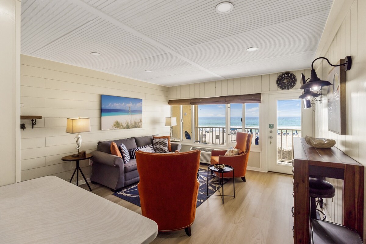 The living room features contemporary furnishings, including a gray sofa and two orange armchairs. A wall of windows offers expansive ocean views and natural light. Decorative elements include a lamp and a coastal-themed artwork, creating a relaxing atmosphere.