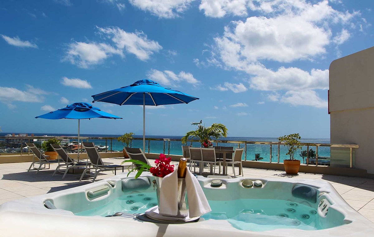 A hot tub is positioned on the terrace, surrounded by vibrant blue umbrellas and lounge chairs. The scenic view showcases the sparkling Caribbean Sea under a bright sky with fluffy clouds, while a dining table is visible in the background.