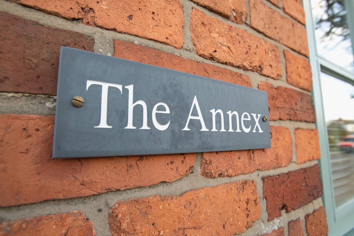 A sign displaying 'The Annex' is mounted on a textured brick wall, featuring a clean, white font. The brickwork exhibits warm tones, and a window can be seen beside the sign.