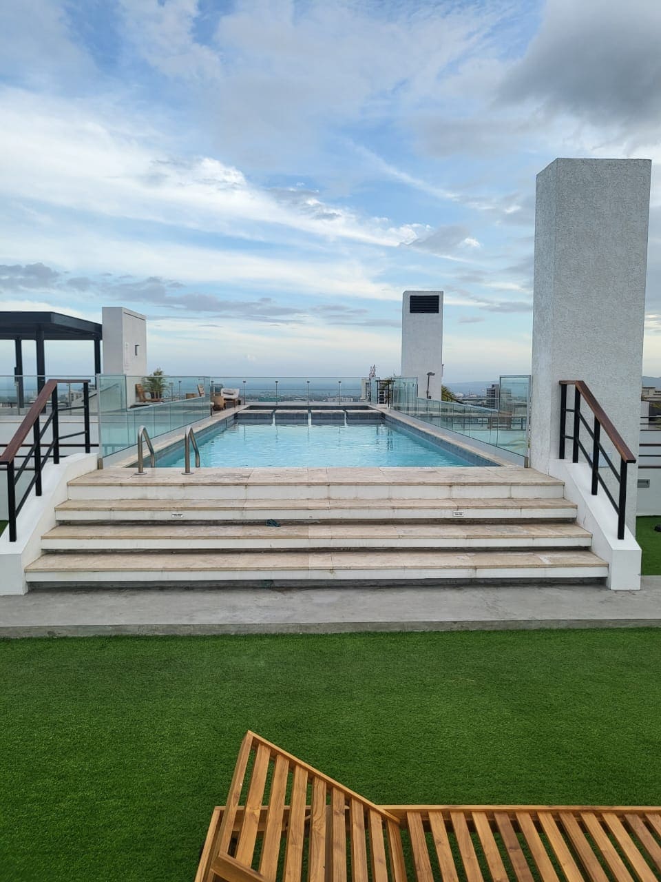 A rooftop pool area is visible, featuring a long pool with steps on one end. Glass railings surround the space, and lounge seating is present on the artificial grass beside the pool. A clear sky and distant views complete the backdrop.
