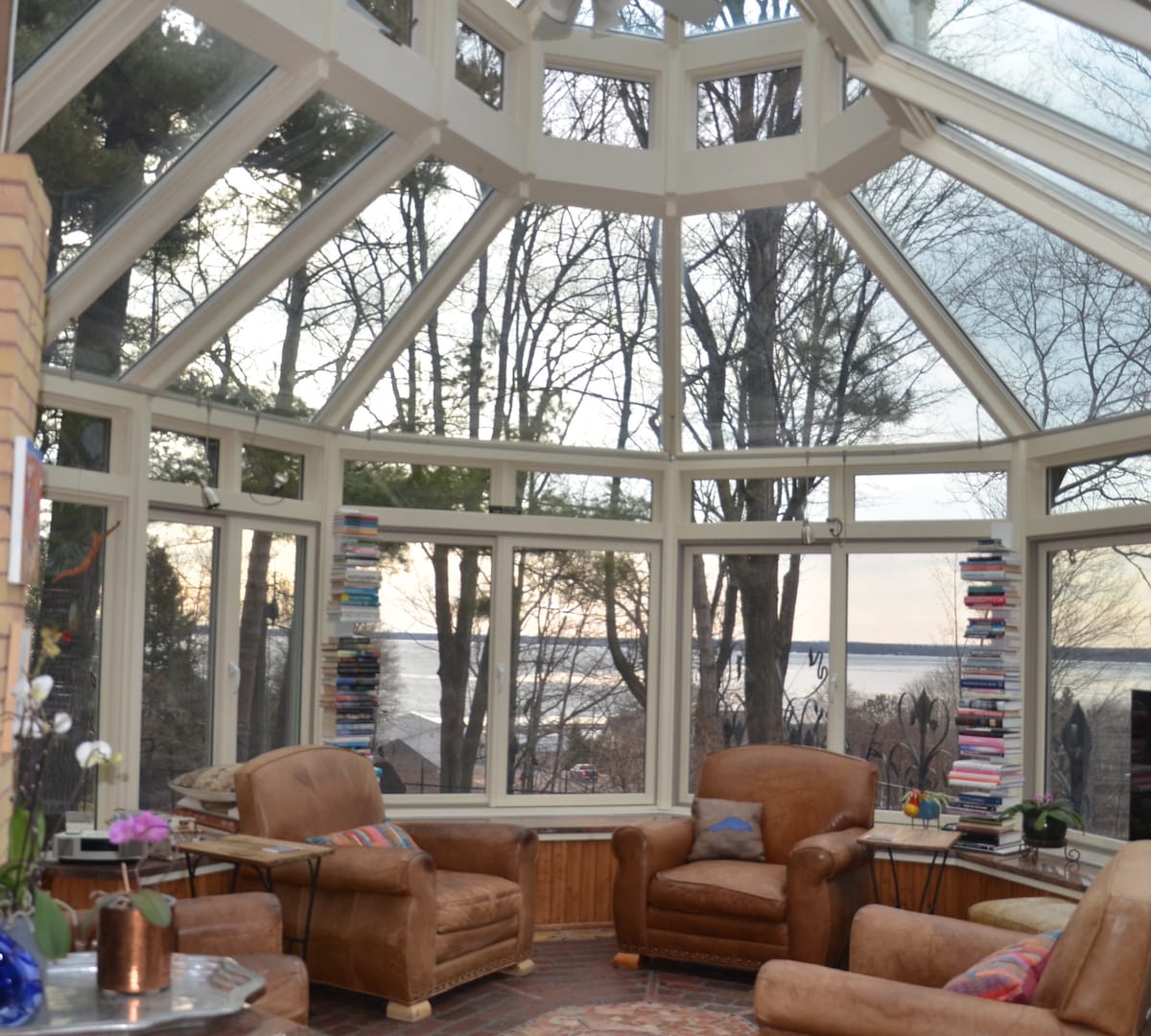 A sunlit solarium features large windows offering panoramic views of the surrounding trees and Lake Superior. Two comfortable brown chairs are positioned beside tables with books. A small plant and flowers add a touch of nature, enhancing the inviting space.