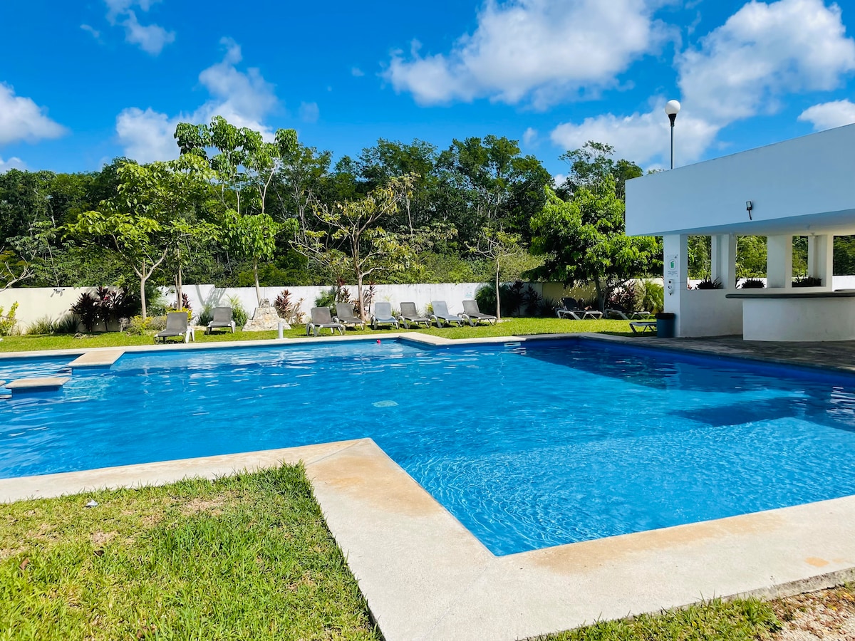 A clear blue swimming pool is surrounded by well-maintained green lawns and trees. Lounge chairs are positioned nearby under a bright sky with scattered clouds. A shaded area with seating is visible, offering a place to relax and enjoy the outdoor space.