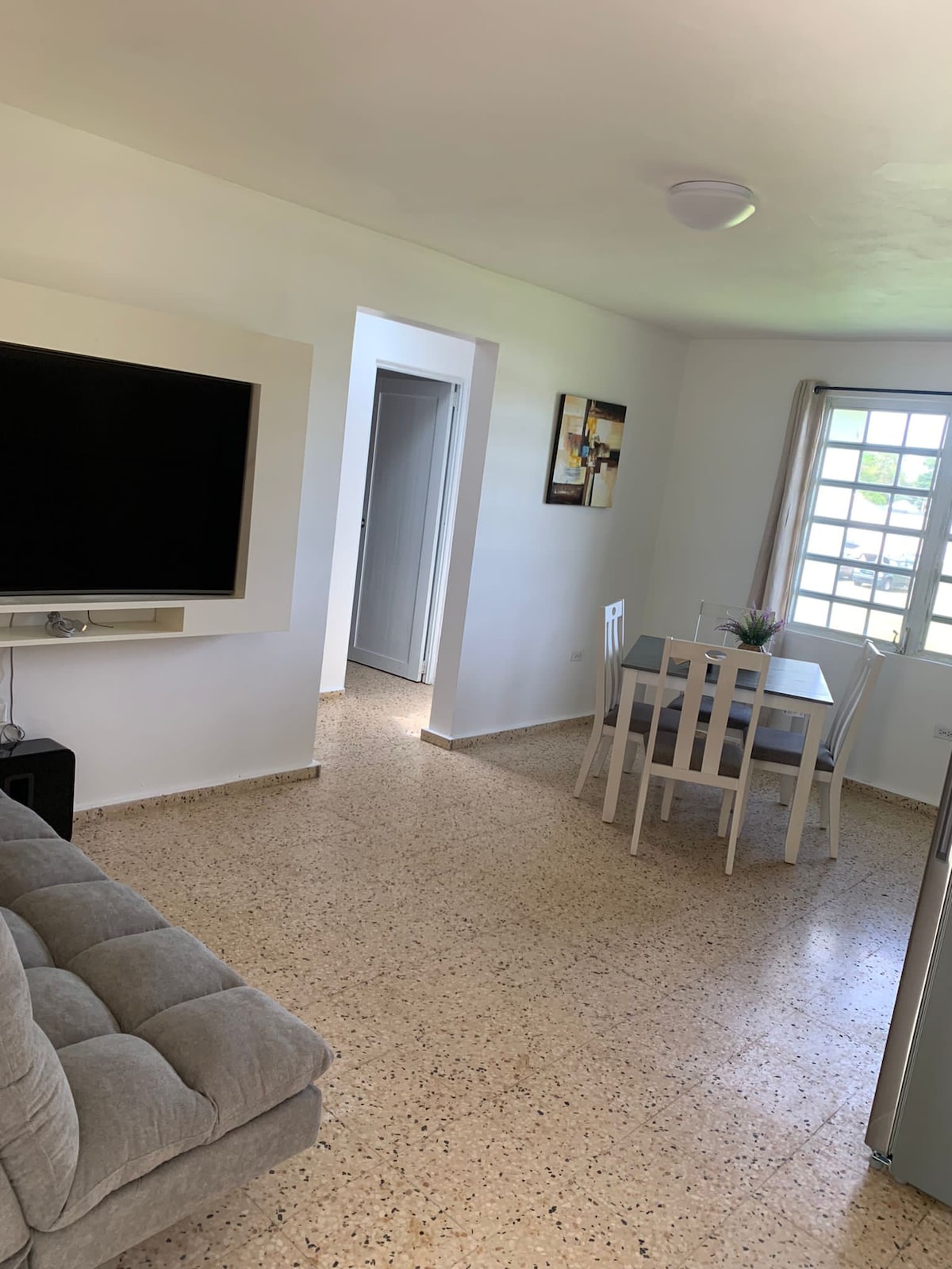 A bright living area features a large flat-screen television mounted on the wall. A small dining table with four chairs is positioned near a window, allowing natural light to fill the space. The floor showcases a light-colored, speckled finish, creating a clean and open atmosphere.
