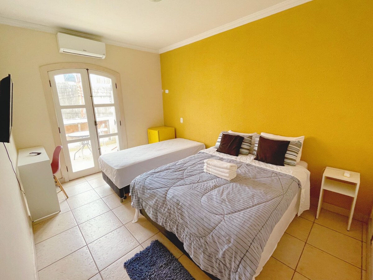 A bright room is presented with a spacious bed and an additional single bed, both neatly arranged. A soft rug complements the tiled floor. Natural light fills the area through large windows, which open to a view outside. A small desk and TV are also visible.
