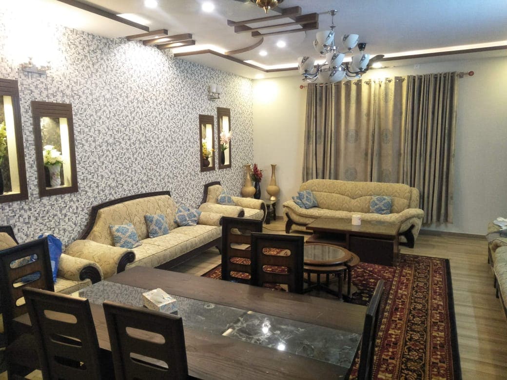 A spacious living area features patterned wallpaper and decorative wall-mounted mirrors. Comfortable seating is arranged around a central wooden table, complemented by a rich area rug. A chandelier illuminates the room, while window treatments add elegance and soften incoming light.