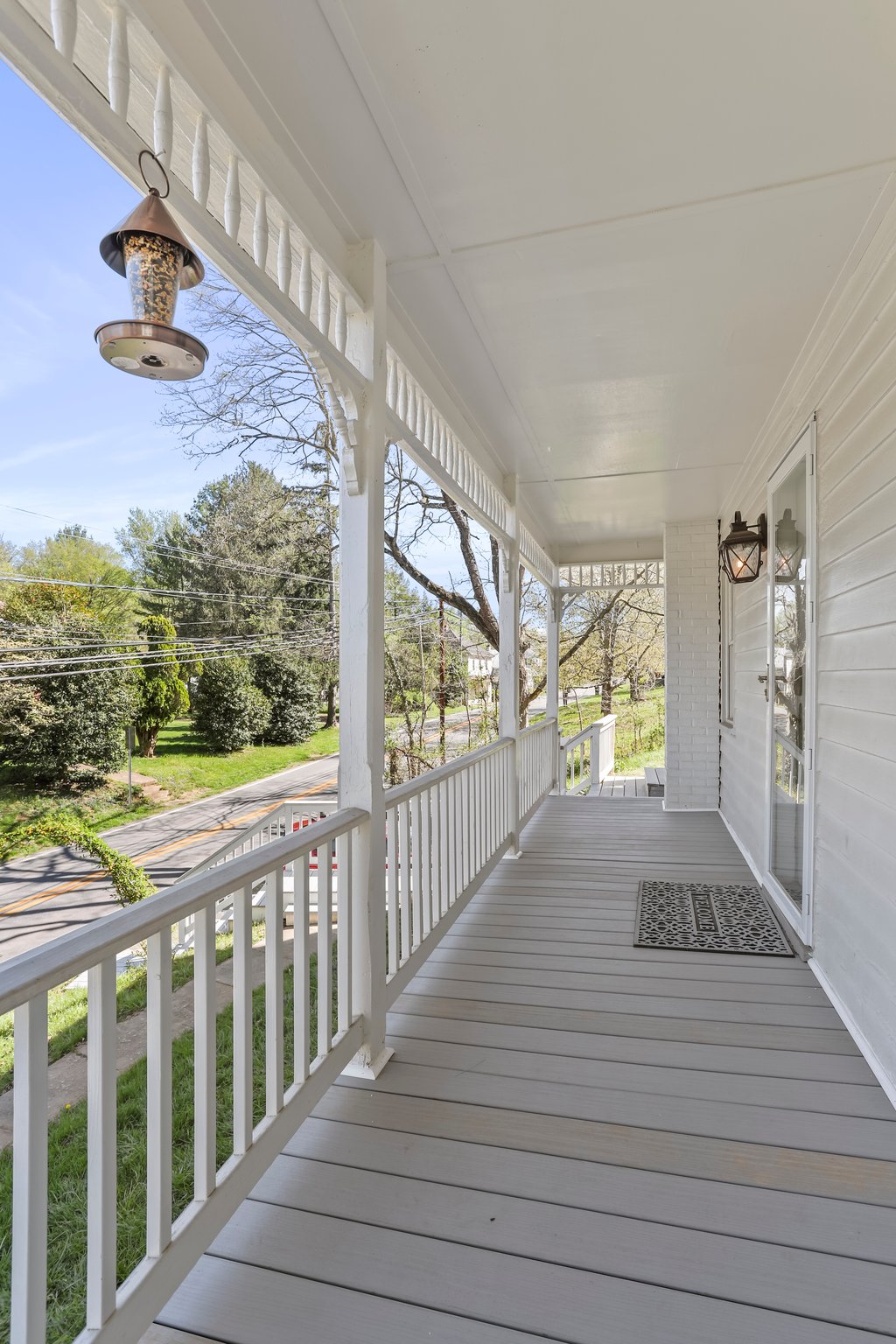A covered porch is shown, featuring white railings and a light-colored floor. The view includes greenery from trees and shrubs along a road. Hanging lanterns are positioned above, providing a sense of welcoming. A textured welcome mat is placed in front of the entrance.