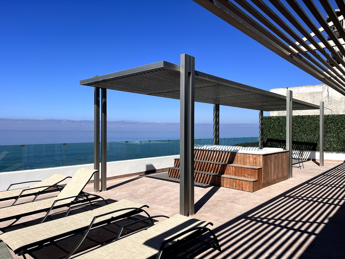 A rooftop area is depicted with a beautiful view of the ocean. A covered jacuzzi sits centrally, surrounded by wooden seating. Several loungers are positioned strategically for relaxation, with a backdrop of a clear blue sky and calm water.