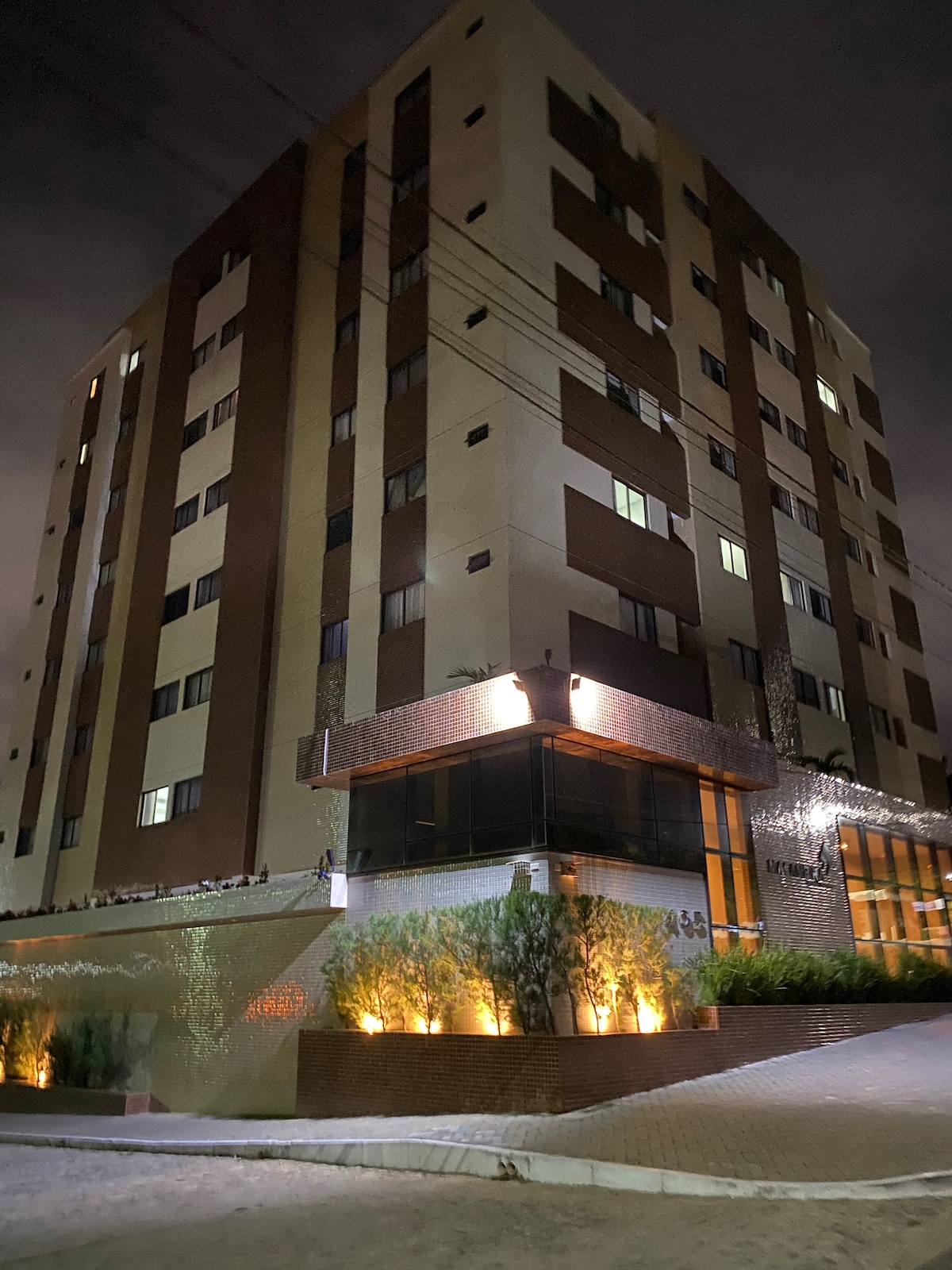 A modern eight-story building is illuminated against the night sky. A combination of brick and neutral panels complements the facade. Soft lighting highlights the entrance area, where greenery is strategically placed along the pathway leading up to the main entrance.