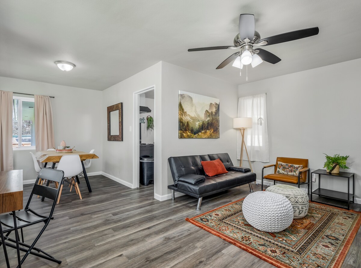 A modern living area features a sleek black sofa, accent chair, and a round pouf. Natural light enters through a window, illuminating the space. A dining table with white chairs is positioned nearby, complemented by stylish decor, including a large artwork and a patterned area rug.