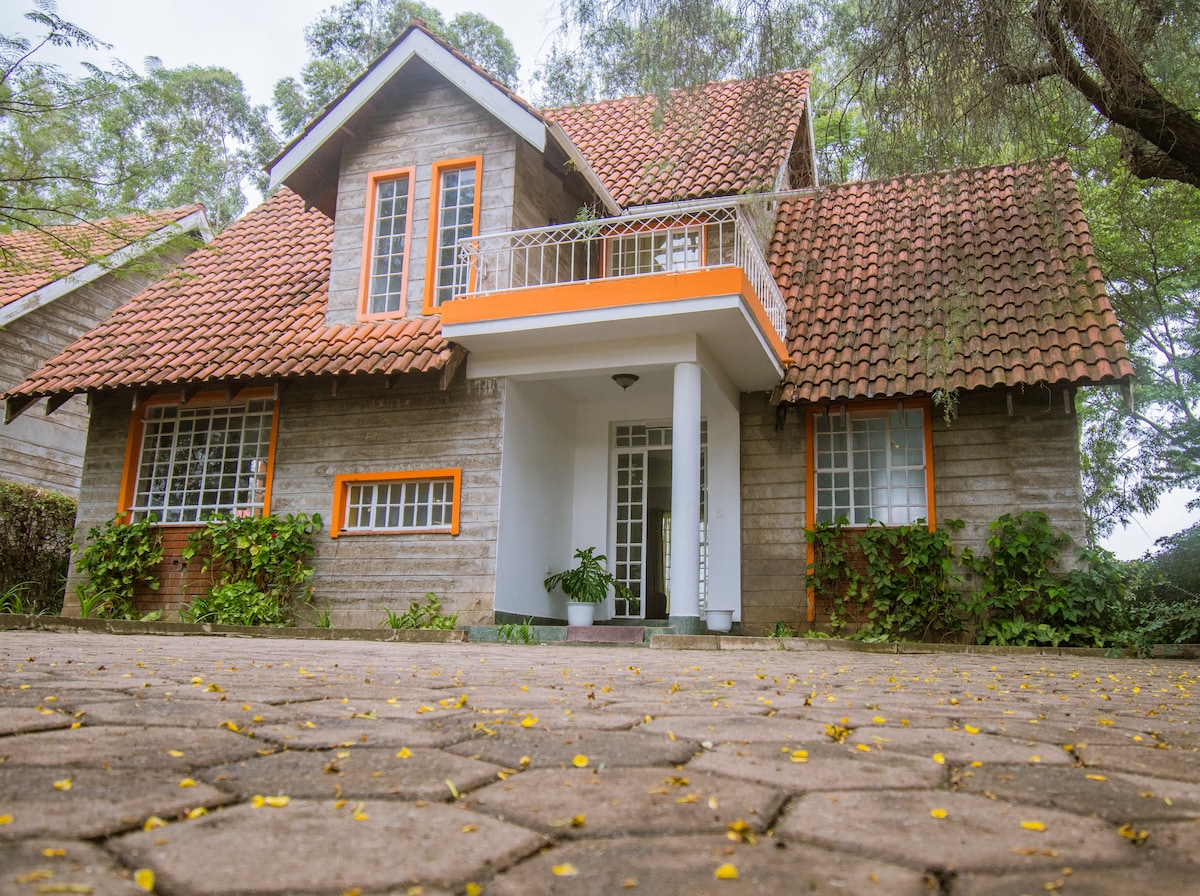 The house features a sloped roof with terracotta tiles and orange accents framing the entrance. A small balcony is positioned above the entrance, surrounded by lush greenery. The pathway leading to the front door is lined with yellow flower petals, enhancing the welcoming atmosphere.