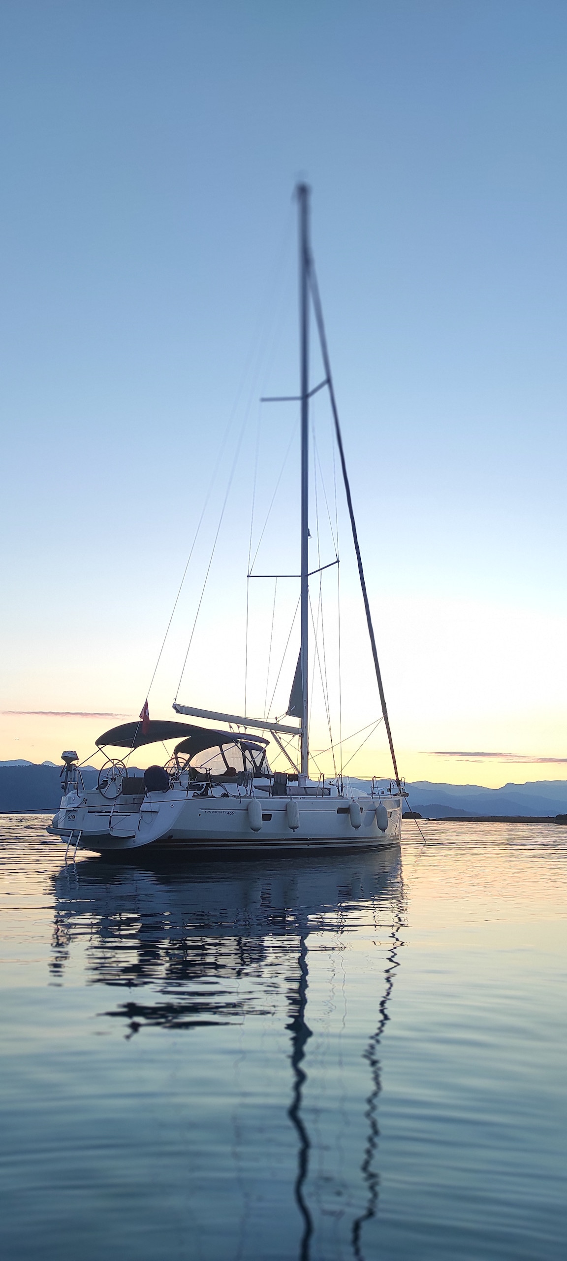 A sailboat is anchored on calm waters, reflecting the soft hues of the twilight sky. The mast rises gracefully above the deck, surrounded by the tranquil scene of nature. The overall atmosphere depicts serenity and a connection to the surrounding waterfront.