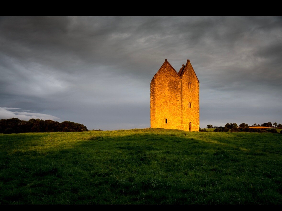 A historic stone structure stands prominently on a grassy hill, bathed in warm light under a dramatic sky. The building features pointed roofs and textured walls, surrounded by open fields, creating a serene and unobstructed landscape.