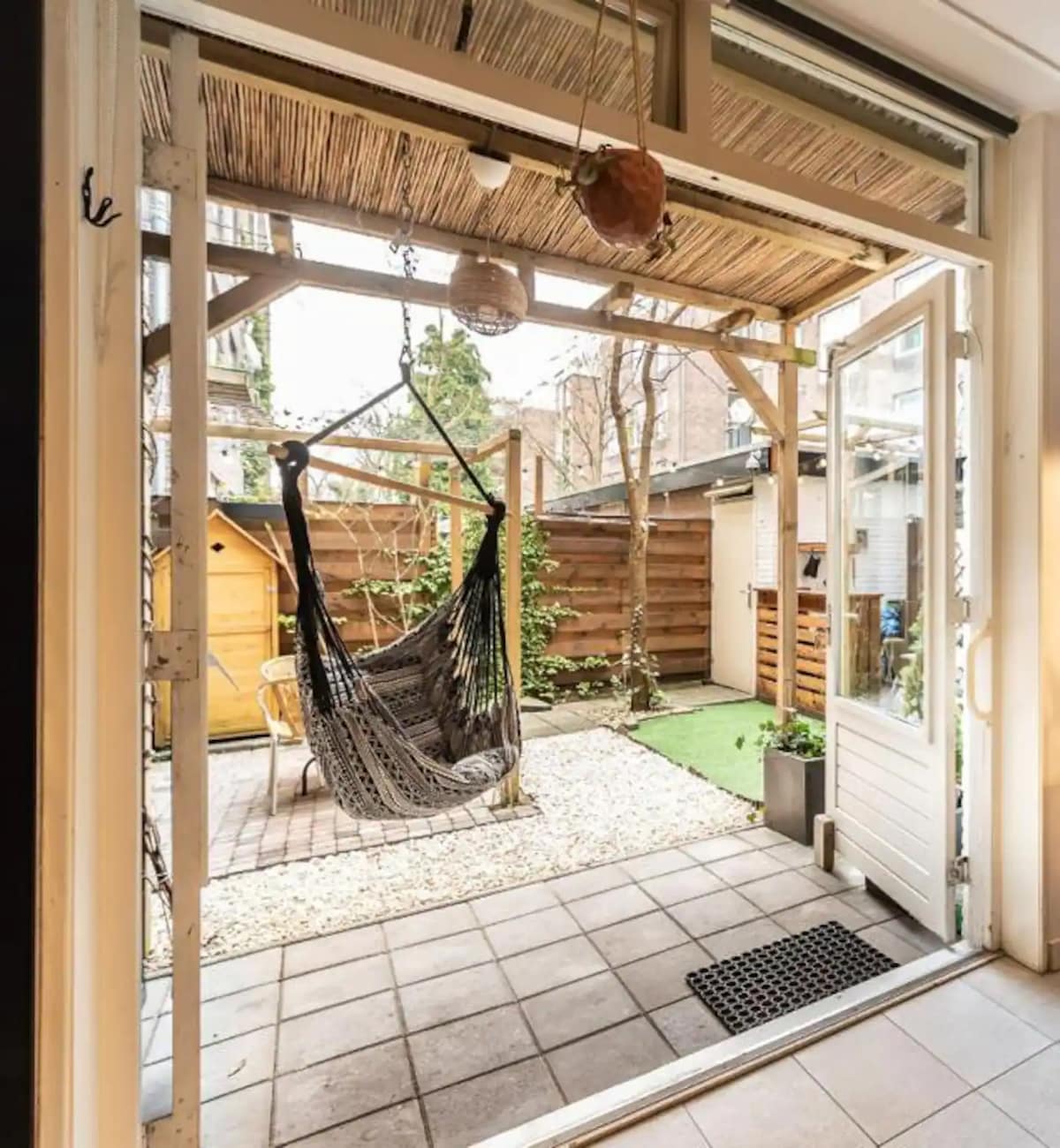 A serene garden space is visible, featuring a hanging hammock suspended from a wooden frame. The area is paved with tiles, surrounded by gravel and greenery. A wooden fence offers privacy, while a wooden structure can be seen in the background.
