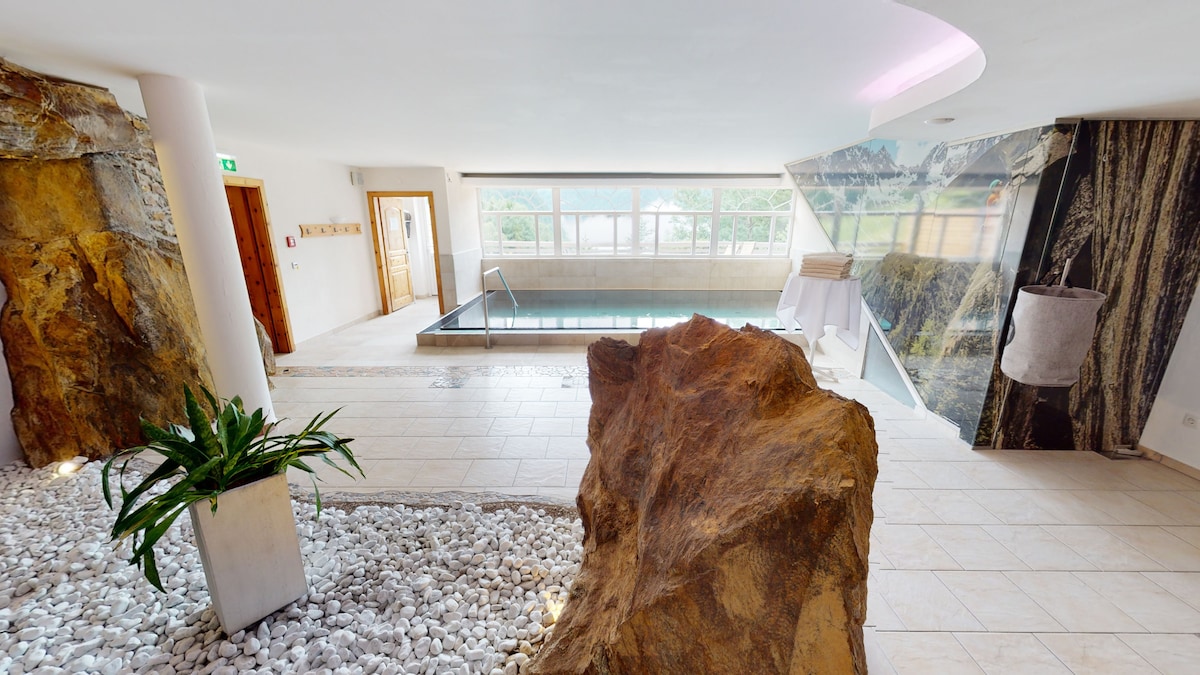 A wellness area features a serene indoor pool surrounded by stone elements. Large windows provide natural light, illuminating the space. Decorative white stones and a green plant add a touch of nature, while elegant marble walls enhance the modern aesthetic.